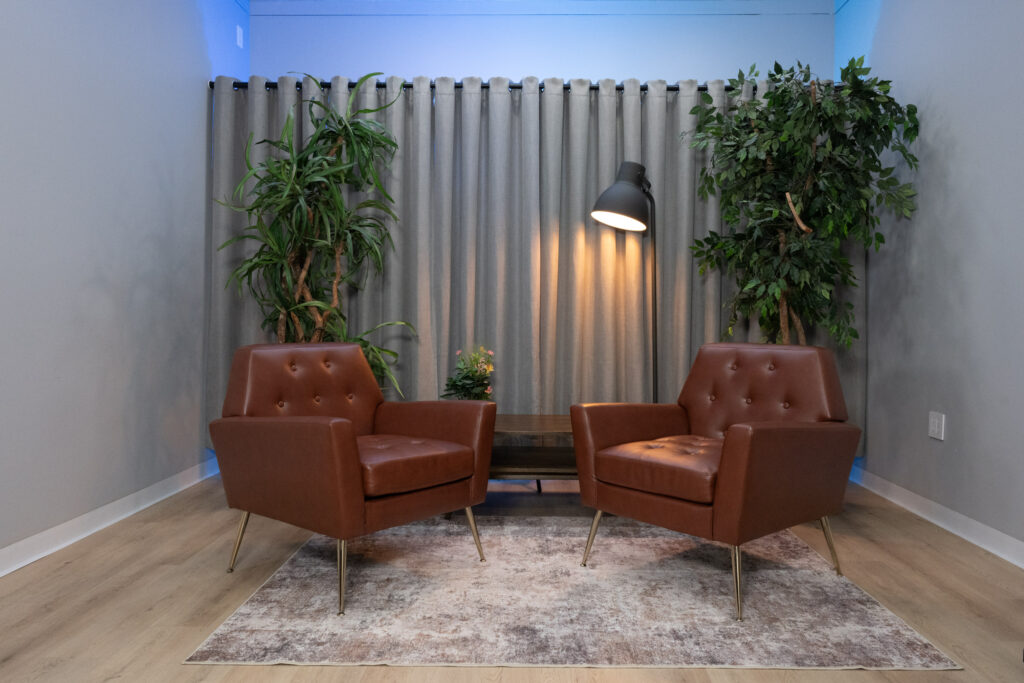 Two tufted brown leather chairs face each other in a soundproofed Houston podcast and video interview studio with warm floor lamp and indoor plants.