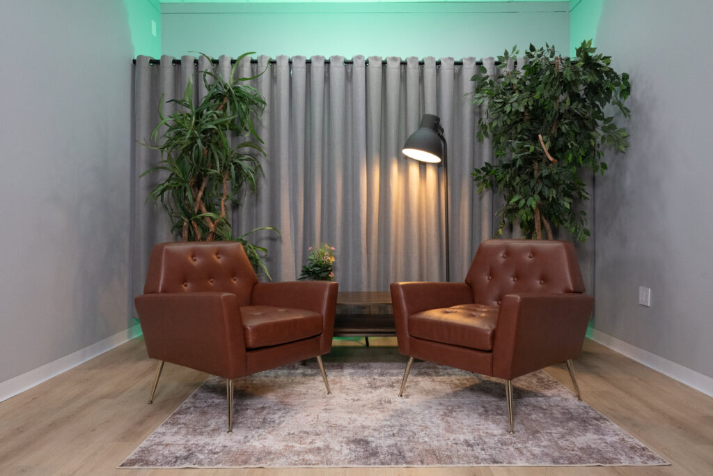 Mid-century leather chairs and indoor plants in a soundproofed Houston podcast and video interview studio with green ambient lighting.