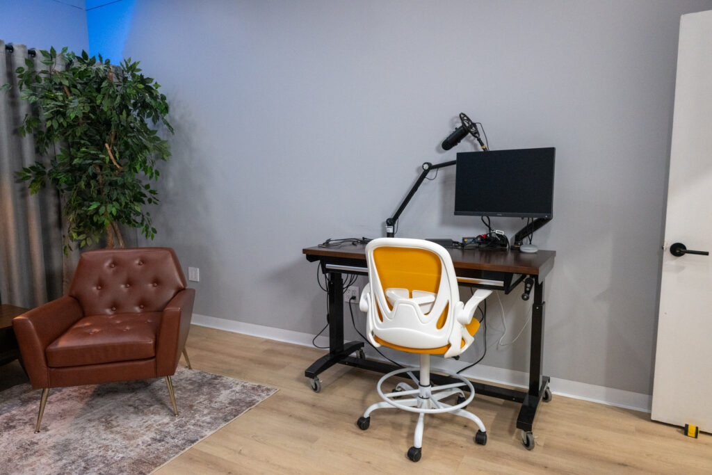 Soundproofed Houston podcast and interview studio with adjustable standing desk, microphone arm, monitor, and orange drafting chair.