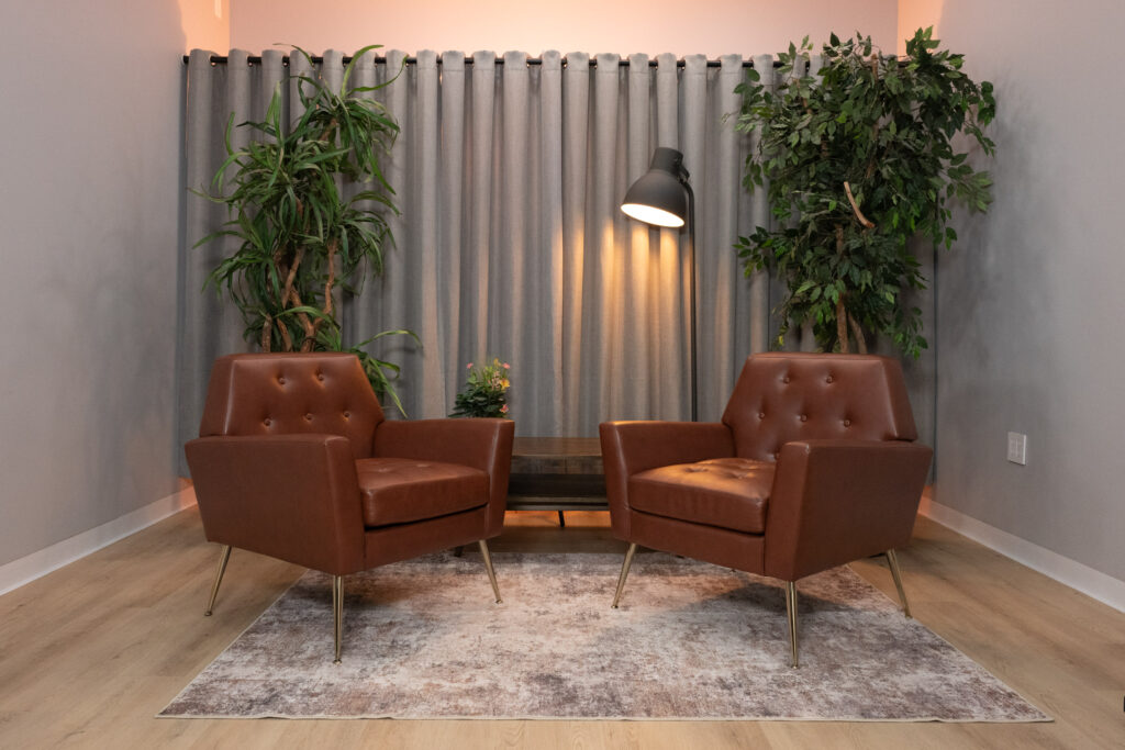 Two brown leather tufted chairs face each other in a warmly lit Houston podcast and video interview studio with plants and floor lamp.